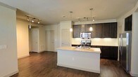 Located at 6040 Parker Blvd in North Richland Hills, Texas, this single-floor residence offers a thoughtfully designed open-concept layout that seamlessly connects the kitchen and living areas. The kitchen is a standout feature, anchored by a large central island with a granite countertop, an integrated sink, and a convenient electrical outlet. Rich, dark-stained cabinetry provides ample storage and creates a bold contrast against the light-toned granite surfaces and classic white tile backsplash. Stainless steel appliances, including a range, microwave, and additional built-in units, complete the modern kitchen setup. Overhead, a combination of track lighting and elegant pendant lights illuminate both the kitchen and the open living space beyond. The living area extends generously from the kitchen, finished with warm, dark hardwood-style flooring that runs throughout the space, adding a cohesive and inviting feel. Neutral wall tones keep the interior bright and versatile. Multiple interior doors hint at well-defined private spaces beyond the main living area, offering a functional and comfortable floor plan suited for everyday living.