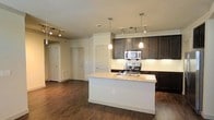 Located at 6040 Parker Blvd in North Richland Hills, Texas, this single-floor residence offers a thoughtfully designed open-concept layout that seamlessly connects the kitchen and living areas. The kitchen is a standout feature, anchored by a large central island with a granite countertop, an integrated sink, and a convenient electrical outlet. Rich, dark-stained cabinetry provides ample storage and creates a bold contrast against the light-toned granite surfaces and classic white tile backsplash. Stainless steel appliances, including a range, microwave, and additional built-in units, complete the modern kitchen setup. Overhead, a combination of track lighting and elegant pendant lights illuminate both the kitchen and the open living space beyond. The living area extends generously from the kitchen, finished with warm, dark hardwood-style flooring that runs throughout the space, adding a cohesive and inviting feel. Neutral wall tones keep the interior bright and versatile. Multiple interior doors hint at well-defined private spaces beyond the main living area, offering a functional and comfortable floor plan suited for everyday living.