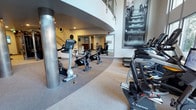 This two-story fitness center offers a well-equipped and thoughtfully designed workout environment. The main floor features an expansive cardio zone lined with treadmills and stationary bikes, including Expresso interactive cycling units and Matrix upright bikes. A dedicated cycling area accommodates several spin bikes, clearly marked with floor indicators for proper spacing. The layout flows naturally, with a curved architectural column and a sweeping staircase adding visual interest to the space.

Large-format black-and-white fitness photography adorns the walls, contributing to an energetic and motivating atmosphere. Full-length mirrors in the cycling area enhance the sense of depth and allow users to monitor their form. The double-height ceiling draws the eye upward to a mezzanine level above, accessible via an interior staircase and enclosed by a glass-panel railing with a warm wood-accented ceiling detail. Natural light filters in through tall windows along the perimeter, brightening the neutral-toned carpet flooring throughout. The overall design balances functionality with a modern, clean aesthetic suited to a serious fitness environment.