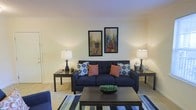 This single-floor residence offers a comfortable and thoughtfully designed living space that balances style with functionality. The living area serves as the heart of the home, featuring warm neutral wall tones that create a bright and inviting atmosphere. Light-colored carpeting adds softness underfoot, complementing the room's overall warmth. A bold navy sofa anchors the seating area, accented by a mix of patterned and solid throw pillows in shades of orange and blue, adding a vibrant pop of color. Flanking the sofa are matching dark wood side tables, each topped with elegant table lamps that cast a warm ambient glow throughout the space. A rectangular wood coffee table sits at the center, grounded by a striped area rug in coordinating tones of blue, cream, and gray. The walls are adorned with a pair of framed abstract cityscape artworks, adding a contemporary artistic touch to the space. The overall ambiance is cozy yet polished, with a careful attention to coordinated décor that makes the living area feel both livable and well-curated. The single-floor layout ensures easy accessibility and a seamless flow throughout the home.