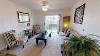 This single-floor residence offers a welcoming and well-appointed living space designed for comfort and ease of living. The living room serves as the heart of the home, featuring plush wall-to-wall carpeting, smooth white walls, and a ceiling fan with integrated lighting that ensures year-round comfort. Natural light pours in generously through a large sliding glass door, which opens directly onto a private outdoor patio — an ideal spot for relaxing or entertaining. The patio provides views of the surrounding exterior landscape, adding a sense of openness to the interior. The living area is spacious enough to accommodate a full seating arrangement, including a sofa, accent chairs, and a central coffee table, all flowing naturally around the room. An arched passageway connects the living space to the kitchen and additional rooms beyond, lending an architectural charm to the layout. The kitchen, visible through the archway, features light cabinetry and ample counter space. The open, connected floor plan allows for easy movement between areas, creating a cohesive and functional living environment. Overall, the space presents a clean, neutral palette that provides a versatile and inviting atmosphere throughout.