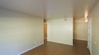 This single-story residence offers a straightforward and functional layout designed for comfortable everyday living. The main living area is finished with warm hardwood-style flooring that extends throughout, complemented by smooth, neutral-toned walls and white baseboards that create a clean, cohesive aesthetic. The space is well-proportioned, providing ample room for a variety of furniture arrangements. The ceiling is smooth and bright, contributing to an airy, open atmosphere. Ventilation is provided through ceiling-mounted air registers, ensuring climate comfort throughout the unit. A hallway visible from the main living area connects to additional rooms, suggesting a practical flow between spaces. A closet opening is also visible, offering convenient built-in storage. The overall condition of the space appears well-maintained and move-in ready, with a neutral palette that provides a versatile backdrop for a range of interior styles. The absence of clutter and the clean finishes throughout give the space a fresh, welcoming feel suited to a variety of living arrangements.