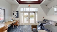 Located at 8732 Town and Country Blvd in Ellicott City, Maryland, this thoughtfully designed community lounge offers a versatile, single-room layout that balances function with comfort. The space is distinguished by its elevated ceiling with a bold, dark-trimmed tray detail that adds architectural character and visual depth throughout.

The room is anchored by a dedicated workspace featuring a long natural wood desk equipped with dual computer monitors and paired with warm cognac leather chairs — ideal for residents seeking a productive environment. Natural light filters through windows fitted with horizontal blinds, keeping the area bright without glare.

At the heart of the lounge, a casual seating arrangement includes a generous curved gray sectional sofa accented with a variety of decorative pillows, accompanied by round dark pedestal tables. Two large rectangular mirrors mounted above the sofa enhance the sense of openness within the space. Patterned dark carpet with geometric line details anchors the room's refined aesthetic.

A set of white-framed French doors with transom windows opens directly onto an outdoor deck, offering views of a pool and landscaped surroundings — seamlessly connecting the interior to outdoor amenity spaces. The open passage to an adjacent room reveals a vaulted ceiling, chandelier, and a reception-style desk, indicating this lounge is part of a larger clubhouse or amenity facility.