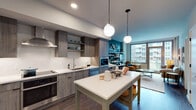 This thoughtfully designed single-floor residence blends contemporary style with functional living. The open-concept kitchen is a standout feature, showcasing wood-grain cabinetry, a textured penny-tile backsplash, white quartz countertops, and stainless steel appliances including a double wall oven and integrated range hood. Globe pendant lights hang above a freestanding island, adding a modern flair while enhancing the workspace. Rich hardwood flooring flows seamlessly from the kitchen into the living area, where large floor-to-ceiling windows and a sliding glass door open to a private balcony, flooding the space with natural light and urban views. A bold teal accent wall anchors the living area, complemented by open shelving for display and a cozy seating nook. The bedroom continues the teal accent wall theme, creating a warm and inviting retreat with ample natural light from a large window. A generously sized walk-in closet offers custom shelving and dual hanging areas for excellent storage. The hallway bathroom features light tile flooring and clean, modern finishes. Throughout, the residence maintains a cohesive contemporary aesthetic, balancing vibrant color choices with neutral tones and quality materials.