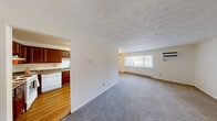 This single-floor, one-bedroom, one-bathroom unit offers a practical and efficient layout across a compact but thoughtfully arranged floor plan. The living and dining areas are open and connected, featuring plush carpet flooring, a textured ceiling, and a large window with vertical blinds that allows natural light to fill the space. A wall-mounted air conditioning unit and baseboard heating provide year-round climate control. A ceiling fan is visible through the pass-through opening from the kitchen, enhancing air circulation in the living area.

The kitchen is separated from the living space by a partial wall with a pass-through opening, creating an accessible, semi-open feel. It features rich, warm-toned wood cabinetry, granite-look countertops, light hardwood-style flooring, and white appliances including a refrigerator and dishwasher. The textured ceiling continues throughout.

The bedroom is generously sized for the overall footprint of the unit, finished with neutral carpet, white walls, a textured ceiling, and baseboard heating. A window provides additional natural light.

The bathroom is clean and bright, featuring white tile walls, a white vanity with an integrated sink, a mirrored medicine cabinet with overhead vanity lighting, and a tiled surround. A hallway connects all rooms efficiently, completing this well-organized layout.