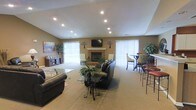 Located at 200 Foxridge Ct in Durham, North Carolina, this single-floor space offers a warm and welcoming atmosphere well-suited for gatherings and everyday use. The main living area features vaulted ceilings with recessed lighting that brightens the room evenly throughout the day. A classic brick fireplace serves as a central focal point, flanked by a wall-mounted flat-screen television and accented with decorative touches that add character to the space. Large windows dressed with vertical blinds allow abundant natural light to fill the interior while offering privacy as needed. The carpeted flooring contributes to a comfortable, casual ambiance throughout. Seating is thoughtfully arranged to accommodate both relaxed lounging and social interaction, with dark leather sofas and a glass-top coffee table anchoring the main seating zone. A dedicated dining area with bar-height table and stools is positioned near the windows, ideal for casual meals or conversation. Potted plants and artwork add life and color to the walls, enhancing the overall inviting feel. The layout is open and functional, making excellent use of the available space to create a cohesive, multi-purpose environment suitable for a variety of activities.