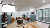 Located at 1400 E Cornwallis Rd in Durham, NC, this community lounge is a well-appointed single-floor space designed for both relaxation and recreation. The room is finished with light wood-tone flooring and a distinctive two-tone wall treatment — crisp white on the upper half and a muted teal-green on the lower — giving the space a modern, cohesive aesthetic. Recessed lighting throughout ensures a bright, welcoming atmosphere, complemented by multiple large windows that invite abundant natural light and views of the surrounding greenery.

The lounge is divided into two functional zones. The seating area features a pair of tufted cognac leather sofas arranged around a round coffee table atop a bold black-and-white checkered rug, alongside a swivel accent chair. A large wall-mounted flat-screen TV is anchored on the teal feature wall above a warm wood media credenza, with an open shelving unit nearby stocked with board games and activities.

The game zone offers a wood-top game table with stools, two classic arcade machines, and a wall-mounted board game panel, creating an engaging entertainment hub. Lush potted plants and framed artwork add finishing touches that bring warmth and personality to this versatile communal space.
