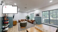 Located at 1400 E Cornwallis Rd in Durham, NC, this community lounge is a well-appointed single-floor space designed for both relaxation and recreation. The room is finished with light wood-tone flooring and a distinctive two-tone wall treatment — crisp white on the upper half and a muted teal-green on the lower — giving the space a modern, cohesive aesthetic. Recessed lighting throughout ensures a bright, welcoming atmosphere, complemented by multiple large windows that invite abundant natural light and views of the surrounding greenery.

The lounge is divided into two functional zones. The seating area features a pair of tufted cognac leather sofas arranged around a round coffee table atop a bold black-and-white checkered rug, alongside a swivel accent chair. A large wall-mounted flat-screen TV is anchored on the teal feature wall above a warm wood media credenza, with an open shelving unit nearby stocked with board games and activities.

The game zone offers a wood-top game table with stools, two classic arcade machines, and a wall-mounted board game panel, creating an engaging entertainment hub. Lush potted plants and framed artwork add finishing touches that bring warmth and personality to this versatile communal space.