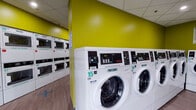 Located at 10 Chestnut Street in Springfield, MA, this commercial laundromat offers a clean, modern, and efficiently organized single-floor facility. The interior is immediately recognizable by its bold yellow-green accent walls, which contrast with the light wood-style flooring and a drop tile ceiling fitted with recessed and flush lighting throughout.

The space is lined with rows of Hercules-branded commercial front-load washing machines and stacked dryer units, all numbered for easy identification. Machines are arranged along the perimeter and in central rows, maximizing the use of floor space while maintaining clear pathways for customers. Stacked dryer units provide additional capacity without consuming extra floor area.

Informational signage, including laundry instruction guides, is posted prominently on the walls to assist users. The facility supports modern, cashless transactions, with posted instructions detailing how to pay using a mobile phone in three easy steps. A folding table is available for customer convenience.

Entry to the space is through a glass-paneled door that allows natural light from a secondary area, adding a sense of openness. Security cameras are visible, ensuring a safe environment. The overall ambiance is bright, functional, and well-maintained, making this laundromat a practical and user-friendly destination for the Springfield community.
