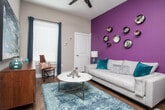 This one-bedroom unit offers a well-designed, open-concept layout that seamlessly connects the kitchen, dining, and living areas. The kitchen is equipped with stainless steel appliances, including a French door refrigerator with an in-door ice and water dispenser, a black electric range, and a built-in microwave. White upper cabinetry contrasts with dark wood-toned lower cabinets, while light quartz countertops and a classic subway tile backsplash add a clean, contemporary finish. A peninsula counter provides additional prep space and casual seating.

The living area is a standout, featuring a bold deep-purple accent wall that adds dramatic visual interest against neutral taupe tones. Wood-style flooring runs throughout the main living areas, adding warmth and cohesion. Natural light filters in through large windows dressed with blinds and curtains.

The bathroom features a quartz-topped vanity with dark wood cabinetry and a framed mirror, connecting conveniently to the bedroom. The bedroom is carpeted and receives ample natural light through large windows. Overall, the unit delivers a modern, energetic aesthetic with thoughtful design details throughout.