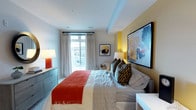 This single-floor bedroom is designed with a bold, contemporary aesthetic that balances comfort and visual interest. The room centers around a generously sized bed dressed in crisp white linens, accented by a vivid red throw blanket and an array of patterned pillows in dark tones, adding texture and depth to the space.

One wall is painted a warm yellow, serving as a striking accent that complements the large, colorful abstract artwork mounted above the bed. A second framed piece adorns the adjacent wall, reinforcing the art-forward design sensibility throughout the room.

Natural light enters through a large window dressed with sheer, floor-length curtains, offering views of the surrounding area while maintaining a bright and airy atmosphere. A red ceramic table lamp on the dresser to one side and a soft bedside lamp on the other provide warm, layered lighting options for the space.

Storage is addressed by a sleek, low-profile dresser positioned near the window, while a clean white surface on the opposite side suggests additional functional furnishing. The overall ambiance is energetic yet comfortable, with the interplay of bold color, curated artwork, and natural light defining the character of this well-designed bedroom.