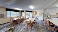 Located at 3185 Karth Rd in White Bear Lake, Minnesota, this single-floor community space is thoughtfully designed to accommodate gatherings, dining, and relaxed socializing. The open layout seamlessly connects multiple functional areas, all unified by patterned carpeted flooring and a neutral warm color palette throughout.

The dining area is generously sized, featuring multiple large tables with wood-framed chairs that can accommodate sizable groups. Large windows dressed with dark valances allow natural light to fill the room while offering views of the surrounding greenery and access to an adjoining outdoor balcony.

The kitchen is well-appointed with crisp white cabinetry, white appliances including a refrigerator with ice maker, stove, microwave, and dishwasher, along with ample counter space. A sideboard with a butcher block top adds additional prep or serving surface, and framed artwork lends a homey touch.

The lounge area provides a comfortable retreat with patterned upholstered armchairs arranged around a white electric fireplace topped with a flat-screen television. French doors with glass panel inserts lead from this sitting area, enhancing the sense of openness. A bookshelf rounds out the space, adding a warm, lived-in character.

Overall, the space offers a welcoming, functional environment well-suited for community events and everyday gatherings.