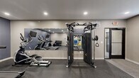 Located at 1633 W Bender Rd in Glendale, Wisconsin, this well-appointed fitness center offers a comprehensive exercise environment within a single, efficiently organized room. The space features rubber flooring throughout the workout area, providing both durability and safety underfoot, while a carpeted rest zone along the windowed wall offers a comfortable area to recover between sets.

The cardio section is anchored by two treadmills and a recumbent bike, each positioned in front of wall-mounted flat-screen televisions set against a bold navy blue accent wall — keeping users entertained during their sessions. A full cable crossover machine serves as the centerpiece of the strength training area, supported by a tiered dumbbell rack with a varied weight selection, a flat weight bench, and a dedicated medicine ball storage rack stocked with multiple sizes.

A large wall mirror spans a significant portion of the room, visually expanding the space and allowing users to monitor their form during workouts. Natural light filters in through multiple windows fitted with white blinds along one wall, and recessed ceiling lighting ensures the room remains bright throughout the day. Two stylish lounge chairs positioned near the windows provide a relaxed seating area. The fitness center is accessible via a glass-paneled entry door, adding a clean, modern touch to the overall design.