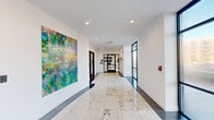 Located at 200–250 Anthony Ave in Mundelein, IL, this single-floor common area presents a polished and contemporary environment spanning a generously sized footprint across three interconnected hallways. The space immediately impresses with its gleaming large-format granite tile flooring, which reflects recessed ceiling lighting throughout, lending the corridors a bright and refined atmosphere.

The hallways serve as the primary circulation and gathering spine of the building, adorned with an eye-catching rotating gallery of large-scale paintings—including vibrant floral abstracts and serene landscape compositions—that add warmth and character to the clean white walls. Floor-to-ceiling black-framed glass windows and entry doors allow abundant natural light to pour in and offer views of the surrounding landscaped exterior.

An elevator lobby anchors one section of the space, featuring dual elevators, a geometric inlaid floor medallion, and classic leather Barcelona-style chairs arranged as a comfortable waiting area, accompanied by additional artwork on the walls. A dedicated mail room offers a practical amenity with built-in mailbox banks flanked by sleek dark paneling and contrasting granite floors. Throughout, the design balances modern minimalism with curated artistic touches, creating a welcoming and functional environment for daily use.