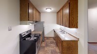 This single-floor residential unit offers a practical and comfortable layout with well-defined living spaces throughout. The open living area is generously sized, featuring neutral-toned carpet, textured ceilings, and a sliding glass door that fills the room with natural light and provides access to an outdoor balcony. A wall-mounted air conditioning unit and baseboard heating ensure year-round comfort. A ceiling fan adds to the airflow in the adjoining area near the kitchen.

The galley-style kitchen is efficiently designed with warm wood cabinetry lining both sides, offering ample storage above and below the counters. It is equipped with a stainless steel refrigerator, an electric range with a ventilation hood, and a double-basin sink set into laminate countertops. Stone-patterned flooring ties the kitchen together with a classic, durable finish.

The bedroom is a calm, carpeted retreat featuring sliding wood-panel closet doors that provide generous storage with hanging space. The warm-toned wood accents on the doors complement the neutral walls and carpeting.

The bathroom is clean and functional, appointed with white square-tile wainscoting, a single-basin vanity with wood cabinetry, and a large wall mirror topped by a multi-bulb vanity light fixture in brushed nickel, providing bright, even illumination.
