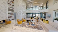 This elegantly designed amenity space makes an immediate impression with its grand, double-height lobby lounge. Soaring ceilings draw the eye upward toward a cascading contemporary chandelier composed of linear LED elements, while a floor-to-ceiling marble feature wall anchors the room with timeless luxury. Polished marble flooring extends throughout, reflecting natural light that pours in through expansive glass walls and doors. The lounge area is furnished with generous cream sectional seating accented by bold gold and black throw pillows, complemented by plush velvet accent chairs in a rich mustard yellow. A glass-railed mezzanine overlooks the space, adding architectural depth and an open, airy ambiance. Built-in bookshelves line one wall, adding a curated, residential warmth to the sophisticated setting.

The game room offers a striking contrast with its crisp white walls and a vibrant red billiards table as the centerpiece. A sculptural globe pendant chandelier provides overhead lighting, while a sleek kitchen bar with red counter stools and stainless steel appliances serves the space with functional style. Large windows bring in abundant natural light.

Completing the amenity suite is a dedicated screening room, outfitted with a large projection screen, recessed lighting, and comfortable carpeted flooring — ideal for private viewings and events.