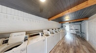 Sugar North Laundromat is a efficiently designed, single-floor commercial laundry facility located in the US. The space is arranged in a long, narrow layout that maximizes functionality while maintaining an open and accessible feel. A row of top-loading commercial washing machines lines one side of the room, complemented by stacked dryer units positioned toward the rear of the space, providing ample laundry capacity for customers. The flooring throughout consists of durable wood-look planks that lend a clean, modern aesthetic to the utilitarian space. Overhead, a distinctive dark-painted shiplap ceiling with warm natural wood exposed beams adds architectural character and visual contrast. The walls feature a mix of painted white brick and textured stucco finishes, contributing to the space's distinctive, rustic-modern appearance. Ceiling fans and flush-mount light fixtures ensure consistent airflow and illumination throughout. Laundry carts are available for customer convenience. A teal entry door at one end provides access, while windows allow natural light to filter into the space. The overall ambiance is clean, practical, and well-maintained, offering a functional environment designed to accommodate the needs of a steady flow of laundry customers.