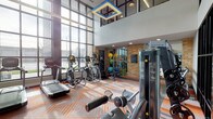 This contemporary fitness center is designed to deliver a premium workout experience within a striking architectural setting. The space is defined by soaring double-height ceilings and expansive floor-to-ceiling windows that line the exterior wall, flooding the interior with abundant natural light and offering open views to the outdoors. An upper-level mezzanine with black-framed glazing adds visual depth and an industrial-modern aesthetic, complemented by warm exposed brick accent walls and a bold geometric-patterned carpet in orange and grey tones.

The cardio zone features a row of Matrix treadmills positioned directly facing the large windows, alongside elliptical trainers and a recumbent bike, allowing users to exercise with an energizing view. A dedicated strength training area houses a comprehensive line of Matrix cable and resistance machines arranged along one wall, with rubber flooring providing safety and durability underfoot. A free weights section offers a full rack of dumbbells and adjustable benches, while a functional training zone is stocked with medicine balls, stability balls, resistance bands, and additional accessories stored on a dedicated rack.

A large flat-screen television mounted on a sleek dark feature wall, paired with a beverage refrigerator, adds convenience and entertainment. Recessed lighting and a distinctive geometric pendant fixture complete the polished, modern ambiance of this well-appointed fitness facility.