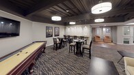 This thoughtfully designed space combines entertainment, recreation, and fitness into a cohesive, stylish layout. The kitchen is a standout feature, outfitted with rich dark cabinetry, light granite countertops, a large central island, stainless steel appliances including double wall ovens and a French door refrigerator, and a mosaic tile backsplash. Industrial-style pendant lights illuminate the workspace, while wood-look tile flooring adds warmth throughout.

Adjacent to the kitchen, the game and lounge area offers a versatile gathering space centered around a classic shuffleboard table. Bistro-height tables and chairs are arranged throughout, complemented by a wall-mounted bar counter with pendant lighting, framed movie posters, and a patterned area rug that anchors the room. A cozy seating nook with a sofa and accent chairs provides a relaxed retreat, and a wall-mounted television adds to the entertainment experience.

The exposed ceiling with dark-painted beams and joists lends an urban, industrial character to the space, while large drum pendant lights provide bright, even illumination. Completing the space is a dedicated fitness room visible through a window partition, equipped with elliptical machines and treadmills. Together, these zones create a dynamic, multi-purpose environment ideal for socializing, recreation, and wellness.