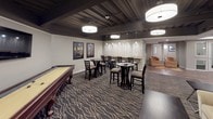 This thoughtfully designed space combines entertainment, recreation, and fitness into a cohesive, stylish layout. The kitchen is a standout feature, outfitted with rich dark cabinetry, light granite countertops, a large central island, stainless steel appliances including double wall ovens and a French door refrigerator, and a mosaic tile backsplash. Industrial-style pendant lights illuminate the workspace, while wood-look tile flooring adds warmth throughout.

Adjacent to the kitchen, the game and lounge area offers a versatile gathering space centered around a classic shuffleboard table. Bistro-height tables and chairs are arranged throughout, complemented by a wall-mounted bar counter with pendant lighting, framed movie posters, and a patterned area rug that anchors the room. A cozy seating nook with a sofa and accent chairs provides a relaxed retreat, and a wall-mounted television adds to the entertainment experience.

The exposed ceiling with dark-painted beams and joists lends an urban, industrial character to the space, while large drum pendant lights provide bright, even illumination. Completing the space is a dedicated fitness room visible through a window partition, equipped with elliptical machines and treadmills. Together, these zones create a dynamic, multi-purpose environment ideal for socializing, recreation, and wellness.
