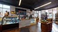 This inviting single-floor specialty shop seamlessly blends a café experience with a curated retail environment. The space is anchored by a stone-clad service counter housing a professional espresso machine, overhead chalkboard menus, and a glass display case showcasing an assortment of prepared foods and confections. A refrigerated display case along the side wall provides additional product storage and visibility. The retail floor features warm hardwood-style flooring throughout, complemented by an exposed dark ceiling with industrial linear lighting that adds an urban, contemporary character. Rustic wooden barrel tables serve as both décor and functional display surfaces, reinforcing an artisanal aesthetic. Floor-to-ceiling curved windows along the front of the space flood the interior with abundant natural light and provide open views to the street, creating an airy, welcoming ambiance. Tall wooden shelving units line the walls, densely stocked with a wide variety of specialty jars, bottles, sauces, and packaged goods. Additional freestanding displays and basket arrangements offer artisan kitchenware and gourmet products. A cozy seating nook nestled within the windowed bay area provides intimate barrel-top tables with folding chairs, ideal for customers to relax. The stone-textured accent walls throughout tie together the warm, rustic-meets-industrial design of this thoughtfully appointed space.