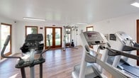 This single-floor fitness center is thoughtfully designed to support a well-rounded exercise routine in a clean, welcoming environment. The space is finished with warm wood-style flooring and crisp white walls, creating a bright and motivating atmosphere. Natural light pours in through multiple large windows and a set of elegant wood-framed French doors that open to an outdoor area, connecting the interior to the surrounding greenery beyond.

The cardio equipment area is anchored by two Life Fitness treadmills positioned side by side, accompanied by additional elliptical trainers and a recumbent stationary bike, offering a full range of low-impact and aerobic options. A multi-functional cable weight machine provides strength training versatility for users of varying fitness levels.

Overhead, flush linear lighting fixtures illuminate the entire space evenly, complementing the natural light throughout the day. A wall-mounted mini-split air conditioning unit ensures a comfortable workout environment year-round. The French doors offer both ventilation and easy access to the outdoors.

The layout is open and uncluttered, allowing ample space to move between machines comfortably. The combination of quality equipment, natural light, and thoughtful finishes makes this fitness center a practical and inviting facility for daily use.