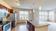 Located at Junction Way in Menomonee Falls, WI, this one-bedroom, one-bathroom unit offers a well-designed layout across a single floor. The open-concept kitchen serves as the centerpiece of the living area, showcasing rich dark wood cabinetry, granite countertops, and a full suite of stainless steel appliances including an electric range, built-in microwave, and refrigerator. A large kitchen island with an integrated sink and pendant lighting above adds both functionality and style. Light hardwood flooring runs throughout the main living and kitchen areas, complemented by multiple windows that invite generous natural light and outdoor views. The bedroom is comfortably proportioned, featuring neutral carpet, a large window overlooking the surrounding landscape, and a dedicated closet for ample storage. The bathroom is cleanly appointed with a dark wood vanity, white countertop, a large mirror, and a full tub-shower combination, all finished with light hardwood-style flooring. Neutral wall tones throughout the unit create a bright, welcoming ambiance, while track lighting in the kitchen enhances both task and ambient illumination. The overall design balances practicality with modern finishes, making it a functional and comfortable living space.