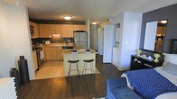 This single-floor residence is designed around an open-concept layout that seamlessly connects the kitchen and living areas, creating a spacious and functional environment. The kitchen is well-appointed with stainless steel appliances, including a refrigerator and range, complemented by warm wood-toned cabinetry and a tile backsplash that adds texture and visual interest. Light stone countertops extend to a central island, which serves as both a prep space and a casual dining area, accommodating two bar-height stools. The tile flooring in the kitchen transitions to rich, dark hardwood-style flooring that flows throughout the remainder of the living space, anchoring the overall aesthetic. A partial wall with built-in storage separates the kitchen zone from an adjacent area, which appears to house laundry appliances, adding practical convenience. The living area is furnished with a plush blue sofa and armchair, creating a comfortable retreat. The open sightlines between spaces enhance the sense of depth and connectivity throughout the unit, while flush ceiling fixtures provide consistent ambient lighting across the interior.