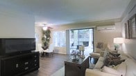 This single-floor residence offers a welcoming and well-designed open-plan layout that seamlessly connects the living and dining areas into one cohesive space. The living area is comfortably appointed, centered around a stylish dark wood coffee table and complemented by neutral-toned seating that creates an inviting atmosphere. A ceiling fan overhead adds both function and a classic touch to the room.

The dining area sits adjacent to the living space, featuring a sleek glass-top table surrounded by white upholstered chairs, making it an elegant spot for everyday meals or entertaining. A wall-mounted flat-screen television and dark media console anchor one side of the room, providing a practical entertainment setup.

Natural light pours generously into the space through large sliding glass doors and wide horizontal blinds, offering direct access to an outdoor patio area with views of lush greenery. A wall-mounted air conditioning unit ensures year-round comfort. The rich, dark hardwood-style flooring runs throughout, grounding the space with warmth and contrast against the light-colored walls and textured ceiling. Decorative touches, including an ornamental mirror and a tall indoor plant, add personality and life to the overall ambiance.
