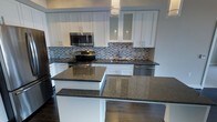 This single-floor space showcases a thoughtfully designed, modern kitchen that serves as its standout feature. The kitchen is outfitted with crisp white shaker-style cabinetry accented by sleek bar-pull hardware, creating a clean and contemporary aesthetic. Rich dark granite countertops provide a striking contrast against the cabinetry and extend across ample workspace, including a generous island or peninsula that offers additional prep and seating opportunities. A mosaic tile backsplash in a blend of grey, black, and white tones adds texture and visual interest to the space, running seamlessly between the upper and lower cabinets. Stainless steel appliances — including a range, over-the-range microwave, and dishwasher — complete the kitchen's polished, functional look. Pendant lighting overhead contributes to the bright, well-lit atmosphere. The dark hardwood flooring grounds the space with warmth and durability, complementing the bold countertops. Upper cabinets with frosted glass panel inserts add an elegant touch, while the overall layout is designed for both efficiency and style, making this kitchen as practical as it is visually impressive.