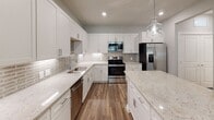 Located at 5401 Memorial Ln in Greensboro, NC, this single-floor residence offers a thoughtfully designed layout with modern finishes throughout. The kitchen is a standout feature, showcasing crisp white shaker cabinetry extending to the ceiling, elegant granite countertops with subtle earth-tone veining, a decorative tile backsplash, and a full suite of stainless steel appliances including a range, microwave, dishwasher, and refrigerator. A large kitchen island provides additional prep and seating space, while a statement glass pendant light adds a stylish touch. Rich wood-look flooring flows through the main living areas, creating a warm and cohesive aesthetic. The living area is generously proportioned with neutral walls and a ceiling fan for comfort. Bedrooms are carpeted for comfort and feature ceiling fans and windows that invite natural light. One bedroom includes built-in shelving, offering versatile storage and display options. A spacious walk-in closet with multi-tiered wire shelving systems provides excellent organization capacity. The bathroom is finished with granite-topped vanity, a tiled tub-shower combination, and stone-look tile flooring, maintaining the home's consistent attention to quality and detail throughout.
