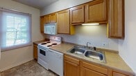 This single-story home at 109 W Colonial Dr in New Castle, Indiana, offers a straightforward, functional layout with several well-defined living spaces throughout. The kitchen is equipped with warm honey-toned wood cabinetry extending to the ceiling, providing generous storage. White appliances, including a coil-top range with overhead microwave, and a double stainless steel sink with laminate countertops complete the practical setup. Tile flooring runs through the kitchen and continues into the full bathroom, which features a vanity with wood cabinetry beneath a white countertop, a wall-mounted light fixture above a large mirror, and a tub-shower combination with chrome fixtures. The living area is spacious and carpeted, with a textured ceiling and a sliding glass door fitted with vertical blinds that allows natural light in and provides direct access to the outdoors, offering pleasant views of surrounding greenery. A pendant light fixture illuminates an adjacent dining or flex area, which also features a pass-through shelf and an additional window. The bedrooms are carpeted, with textured ceilings and windows dressed with blinds, creating calm, neutral spaces adaptable to various uses. Overall, the home presents a clean, neutral palette throughout.