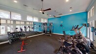 This fitness center is a comprehensively outfitted single-floor facility designed to accommodate a wide range of workout routines. The space is organized into distinct functional zones, each serving a specific training purpose. Bold teal accent walls contrast with crisp white trim and ceilings throughout, creating an energetic and visually cohesive environment. Dark carpeted flooring runs across the facility, providing a durable and practical surface underfoot.

The main workout area houses a robust selection of strength training equipment, including a multi-station cable machine, a rack of dumbbells in graduated weights, and a flat weight bench. Ceiling fans are mounted overhead to maintain air circulation during intense sessions, while recessed lighting ensures consistent illumination throughout. Full-length mirrors line portions of the walls, visually expanding the space and allowing users to monitor their form.

A dedicated cardio zone positioned along a wall of large floor-to-ceiling windows features elliptical machines, Spinner cycling bikes, and a recumbent bike, benefiting from generous natural light and outdoor views. A flat-screen television is wall-mounted for entertainment during workouts.

A separate alcove area stores stability balls, medicine balls in varying sizes, and exercise mats, providing ample resources for stretching and functional training exercises.