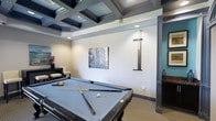 This single-floor space is designed as an upscale recreational and social area, centered around a full-sized billiards table finished in sleek gray felt. The table is prominently positioned beneath a striking coffered ceiling, which serves as the room's defining architectural feature. The ceiling's deep, geometric box moldings are finished in dark charcoal tones, creating a bold contrast against the lighter walls and adding considerable visual depth to the space.

Recessed lighting is integrated into the coffered ceiling, providing even, ambient illumination throughout the room. A wall-mounted cue rack keeps billiards equipment neatly organized and within easy reach, while framed wall art adds a decorative touch to the clean, white walls. Decorative columns with dark trim echo the ceiling's color palette and reinforce the room's refined aesthetic.

Adjacent to the billiards area, a secondary seating or lounge zone is visible, furnished with a cushioned bench and accented with fresh florals, offering a comfortable space for spectators. An accent wall in soft teal adds a subtle pop of color. Overall, the space exudes a polished, club-like atmosphere suited for leisure and entertainment.