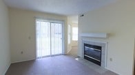 This single-floor space offers a welcoming and functional layout designed for comfortable everyday living. The main living area is a standout, centered around a classic tiled fireplace with a clean white mantel that serves as the room's focal point. Soft, plush wall-to-wall carpeting runs throughout, adding warmth and comfort underfoot.

Natural light is a defining feature of this space, streaming in generously through a full-width sliding glass door fitted with vertical blinds, offering easy access to the outdoors and a seamless connection to the surrounding greenery visible through the glass. An additional window further brightens the interior, creating an airy, open atmosphere.

The walls are finished in a soft, neutral tone that complements the light-filled environment and provides a versatile backdrop for a variety of interior styles. A textured ceiling adds subtle architectural character throughout. Practical details such as a ceiling vent and recessed architectural transitions between spaces reflect a thoughtful, well-considered layout. Overall, the space exudes a calm, inviting ambiance that balances simplicity with comfort.