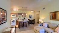 This single-floor residence offers a thoughtfully designed open-concept layout that seamlessly connects the kitchen, dining, and living areas into one cohesive, welcoming environment. The kitchen is well-appointed with warm wood cabinetry, a built-in wine rack, granite countertops, and a breakfast bar with bar seating — ideal for casual dining or entertaining. White appliances, including a range and microwave, complement the classic cabinetry, while track lighting illuminates the workspace effectively. Adjacent to the kitchen, the dining area comfortably accommodates a full table and chair set, making it a natural gathering space. The living area is carpeted and bathed in warm ambient light from a ceiling fan fixture, creating a relaxed and comfortable atmosphere. Neutral, soft tones carry throughout the space, lending a calm and inviting feel. A hallway visible from the main living area suggests additional private rooms beyond, including what appears to be a bedroom. The overall design balances functionality with comfort, making efficient use of the open floor plan while maintaining a warm, residential ambiance throughout.