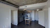 This single-floor space showcases a compelling blend of industrial and contemporary design. The open-concept kitchen is a standout feature, outfitted with sleek dark cabinetry, stainless steel appliances including a French door refrigerator and gas range, and a striking mosaic tile backsplash that adds texture and visual interest. A central kitchen island with a stainless steel top provides additional workspace and casual dining potential. Overhead, an exposed industrial ventilation duct runs prominently across the raw concrete ceiling, lending the space an urban loft character that contrasts beautifully with the refined finishes below. Track lighting illuminates the kitchen area, while flush-mount fixtures provide ambient light throughout. Rich, dark hardwood flooring runs seamlessly across the space, grounding the design with warmth and continuity. Multiple doorways lead to additional rooms, suggesting a well-connected and functional floor plan. The overall ambiance is modern and sophisticated, with a cool, neutral color palette of grays and deep charcoals that gives the space a polished, urban aesthetic well-suited to contemporary living.