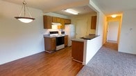 This single-floor residential unit offers a practical and functional layout designed for comfortable everyday living. The kitchen is equipped with warm-toned wooden cabinetry, dark countertops, a built-in sink, a range, a microwave, and a refrigerator. It flows seamlessly into the living area via a peninsula-style breakfast bar, creating an open-concept feel that encourages easy interaction between spaces. The kitchen features durable hardwood-style flooring, while the adjacent living area is finished with plush carpeting throughout. The living room is bright and airy, benefiting from a large sliding glass door that opens onto a private balcony with outdoor views of surrounding greenery, allowing natural light to fill the space. The bedroom is a well-proportioned, carpeted room featuring a window that frames pleasant outdoor views and provides ample natural light. The full bathroom includes a single-basin vanity with wooden cabinetry and a grey countertop, a toilet, and a tub-shower combination with white tile surround, all finished with neutral, easy-to-maintain tile flooring. The overall aesthetic of the unit is clean and neutral, with white walls throughout that offer a versatile backdrop for any style of furnishing.