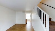This two-story space offers a straightforward and functional layout designed for comfortable everyday living. The main level is anchored by an open living area featuring warm hardwood-style flooring that extends throughout, contributing a sense of continuity and warmth. Crisp white walls and a smooth white ceiling create a clean, neutral backdrop that enhances the brightness of the space. A classic white staircase with dark balusters is prominently positioned along one side of the living area, providing an elegant transition to the upper floor while adding a timeless architectural detail. Natural light filters in from a window visible at the far end of the main level, illuminating the open passage that connects the living area to an adjacent room. A ceiling vent ensures climate control throughout the space. The connected area, partially visible through the open doorway, continues the same hardwood-style flooring and hints at additional functional space, possibly a kitchen or entryway, further reinforcing the cohesive design. The overall ambiance is bright, airy, and move-in ready, with a neutral palette that offers flexibility for a variety of interior styles and furnishing choices.