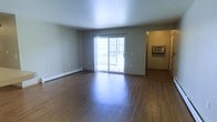 This single-floor space centers around an open living area finished with smooth, medium-toned hardwood-style flooring that extends throughout, reflecting natural light and lending warmth to the interior. The room is defined by neutral gray walls and a plain white ceiling, creating a clean and understated aesthetic well-suited for a variety of furnishing styles.

A sliding glass door positioned along the far wall serves as the primary source of natural light, offering a view onto an outdoor balcony. The door allows daylight to fill the space and provides convenient outdoor access. A baseboard heating unit runs along the lower portion of the adjacent wall, indicating a practical in-unit heating system.

To the right, a hallway leads to additional areas of the unit. Visible within the hallway is a wall-mounted air conditioning unit, providing supplemental climate control. The transition between spaces is framed by warm-toned door casings and baseboards that complement the flooring throughout.

Overall, the space presents a functional and straightforward layout with durable finishes, practical climate control features, and a comfortable level of natural light.