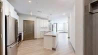 This contemporary single-floor residence showcases a clean, modern design with a neutral color palette and an open, airy layout. The heart of the home is a well-appointed kitchen featuring crisp white cabinetry, light granite countertops, a large center island with an undermount sink, and a full suite of stainless steel appliances including a refrigerator, electric range, and over-the-range microwave. Elegant globe pendant lights hang above the island, while a built-in wine cooler and open shelving add functional character. Light wood-look flooring flows seamlessly throughout the main living areas, complementing the bright, open-concept design that connects the kitchen to the living space. The living area is bathed in natural light through large windows fitted with horizontal blinds, and a ceiling fan adds comfort to the space. The bedrooms offer plush carpeting, multiple windows for abundant natural light, and generous proportions. A well-designed bathroom includes a granite-topped vanity, a large mirror, and direct access to a walk-in closet with wire shelving organization systems. Throughout the residence, taupe-toned interior doors with paneled detailing provide a refined contrast to the white walls and light finishes, creating a cohesive and polished aesthetic.