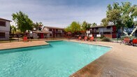 Located at 2025 W Indian School Rd in Phoenix, Arizona, this outdoor amenity space centers around a generously sized swimming pool with crystal-clear turquoise water. The pool is framed by a spacious concrete deck that provides ample room for relaxation and recreation. Bold red lounge chairs and patio furniture with red umbrellas are arranged throughout the pool deck, creating a vibrant, resort-inspired atmosphere well-suited to Arizona's sunny climate. The entire area is enclosed by a black metal fence, providing both security and a defined sense of space. Mature trees, including palm trees and large shade trees, are positioned throughout the surrounding grounds, offering natural shade and a lush visual contrast against the desert setting. The courtyard-style layout is flanked on multiple sides by two-story residential buildings featuring a distinctive red and gray exterior with red accent doors, accessible via exterior stairways and walkways. The combination of the vivid color palette, mature landscaping, and well-maintained pool deck creates an inviting and energetic communal outdoor environment that is characteristic of Phoenix living.