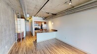 Located in Denver, CO, this single-floor urban loft delivers a compelling blend of industrial character and modern comfort. The open-concept living and kitchen area is defined by soaring exposed concrete ceilings with visible ductwork and conduit, lending an authentic industrial aesthetic. Track lighting and elegant pendant lights illuminate the space, highlighting the warm wood-look flooring that runs throughout.

The kitchen is well-appointed with stainless steel appliances — including a refrigerator, range, and microwave — complemented by warm wood cabinetry, a built-in wine rack, and granite countertops. A peninsula with a stainless steel sink and matching granite surface provides both additional prep space and a natural divide between the kitchen and the open living area.

A standout feature of the living space is a dramatic exposed concrete accent wall, adding raw texture and visual depth to the room, while exposed ductwork overhead reinforces the loft's industrial design language.

The bathroom is finished with tile flooring, a granite-topped wood vanity, and a glass-enclosed shower, offering a clean and functional design. The overall layout flows efficiently, with a long corridor connecting the living spaces to private areas. This Denver loft is an ideal space for those who appreciate urban industrial design paired with quality modern finishes.