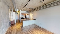 Located in Denver, CO, this single-floor urban loft delivers a compelling blend of industrial character and modern comfort. The open-concept living and kitchen area is defined by soaring exposed concrete ceilings with visible ductwork and conduit, lending an authentic industrial aesthetic. Track lighting and elegant pendant lights illuminate the space, highlighting the warm wood-look flooring that runs throughout.

The kitchen is well-appointed with stainless steel appliances — including a refrigerator, range, and microwave — complemented by warm wood cabinetry, a built-in wine rack, and granite countertops. A peninsula with a stainless steel sink and matching granite surface provides both additional prep space and a natural divide between the kitchen and the open living area.

A standout feature of the living space is a dramatic exposed concrete accent wall, adding raw texture and visual depth to the room, while exposed ductwork overhead reinforces the loft's industrial design language.

The bathroom is finished with tile flooring, a granite-topped wood vanity, and a glass-enclosed shower, offering a clean and functional design. The overall layout flows efficiently, with a long corridor connecting the living spaces to private areas. This Denver loft is an ideal space for those who appreciate urban industrial design paired with quality modern finishes.