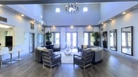 This two-story space makes an immediate impression with its grand, open lobby area that showcases soaring ceilings and an abundance of natural light streaming through large glass doors and strategically placed upper transom windows. A striking multi-arm chandelier serves as a focal centerpiece overhead, complementing the warm recessed lighting that illuminates the space throughout.

The main gathering area is thoughtfully arranged with a curated seating ensemble, featuring plush upholstered sofas and sleek armchairs with metallic frames positioned around a glass-top coffee table, all anchored by a soft area rug atop rich, dark hardwood flooring. Floor-to-ceiling white drape curtains frame the expansive glass entryway, adding an elegant, airy quality to the room.

Lush potted tropical plants are symmetrically placed throughout, introducing vibrant greenery that enlivens the contemporary aesthetic. The walls feature tasteful artwork, and the overall color palette of cool grays, whites, and deep tones creates a refined, modern ambiance. The two-floor layout further enhances the sense of volume and grandeur, making this a welcoming and visually impressive environment suited for both gathering and reception purposes.