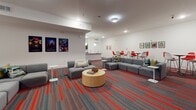 Located at 2008 Third Ave S in Birmingham, Alabama, this single-floor communal space combines modern design with functional versatility. The main lounge area is an expansive, open-plan room anchored by a striking feature wall adorned with a circuit board-inspired graphic mural in red and grey, against which a wall-mounted flat-screen television is centered. The bold aesthetic is carried throughout by vibrant red-striped carpeting that energizes the space and ties the design together cohesively.

Flexible seating arrangements accommodate a variety of uses, from casual socializing to group gatherings. Multiple modular grey sofas are arranged around low side tables and a cylindrical wooden coffee table, while an assortment of round high-top and standard dining tables paired with colorful chairs — in both red and chartreuse — provide additional options for dining or collaborative work. Framed artwork and posters are displayed throughout, lending a curated, creative atmosphere to the space.

Adjacent to the lounge, a well-appointed kitchen features dark espresso cabinetry, granite countertops, a subway tile backsplash, stainless steel sinks, and light-toned tile flooring. Globe pendant lights illuminate the workspace, making it well-suited for catering or event preparation. Together, these areas form a welcoming, design-forward environment ideal for community events and social gatherings.