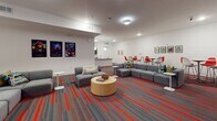 Located at 2008 Third Ave S in Birmingham, Alabama, this single-floor communal space combines modern design with functional versatility. The main lounge area is an expansive, open-plan room anchored by a striking feature wall adorned with a circuit board-inspired graphic mural in red and grey, against which a wall-mounted flat-screen television is centered. The bold aesthetic is carried throughout by vibrant red-striped carpeting that energizes the space and ties the design together cohesively.

Flexible seating arrangements accommodate a variety of uses, from casual socializing to group gatherings. Multiple modular grey sofas are arranged around low side tables and a cylindrical wooden coffee table, while an assortment of round high-top and standard dining tables paired with colorful chairs — in both red and chartreuse — provide additional options for dining or collaborative work. Framed artwork and posters are displayed throughout, lending a curated, creative atmosphere to the space.

Adjacent to the lounge, a well-appointed kitchen features dark espresso cabinetry, granite countertops, a subway tile backsplash, stainless steel sinks, and light-toned tile flooring. Globe pendant lights illuminate the workspace, making it well-suited for catering or event preparation. Together, these areas form a welcoming, design-forward environment ideal for community events and social gatherings.