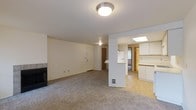 This single-floor residence offers a clean, functional layout designed to maximize livability. The main living area is finished with wall-to-wall carpeting and anchored by a fireplace, creating a cozy focal point for the room. Neutral white walls and flush ceiling lighting contribute to a bright, understated ambiance throughout the space. A half-wall partition subtly defines the transition between the living area and the rest of the home, maintaining an open and airy feel without fully closing off the floor plan. Beyond the partition, the kitchen is a standout feature, benefiting from a skylight that bathes the space in natural light. White cabinetry lines the walls, offering generous storage, while light-toned countertops and flooring give the kitchen a clean, practical character. A peninsula-style counter provides additional workspace and a casual dining option. The entryway is clearly defined, with a front door visible from the living room, lending a straightforward and accessible flow to the overall floor plan. Additional interior doors suggest dedicated spaces for a hallway or utility area, rounding out this well-organized single-level home.