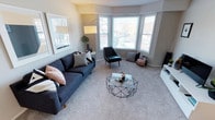 This single-floor space offers a thoughtfully designed and welcoming interior with a modern aesthetic. The standout feature of the living area is a graceful bay window that floods the room with abundant natural light, framing pleasant outdoor views and creating an airy, open atmosphere. Soft, plush carpeting runs throughout the living space, adding warmth and comfort underfoot. The living room is generously proportioned, offering ample space for relaxed seating arrangements. A deep navy sofa anchors the room alongside a sleek geometric glass-top coffee table, a stylish accent chair, and a contemporary arc floor lamp that adds a refined touch. Neutral warm-toned walls provide a versatile backdrop that complements both the furnishings and the natural light pouring in through the bay windows. The overall ambiance is clean, modern, and comfortable — a space that balances style with livability. The open layout encourages easy movement and a sense of spaciousness, making the living area feel both functional and inviting. Subtle décor accents, including a large framed wall print and a potted plant, contribute to the space's polished yet approachable character.