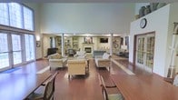 This single-floor communal space exudes a warm and inviting atmosphere, thoughtfully designed to encourage relaxation and social interaction. The expansive main lounge area is anchored by a classic fireplace framed with a clean white mantel, flanked on either side by built-in bookshelves that lend a refined, library-like character to the room. Soaring ceilings supported by elegant columns amplify the sense of openness, while rich hardwood flooring flows throughout, adding warmth and cohesion to the space. Multiple seating arrangements — featuring upholstered armchairs and sofas in neutral, traditional patterns — are thoughtfully positioned to create intimate conversation areas within the larger open plan. A wall-mounted flat-screen television above the fireplace provides an entertainment focal point. Natural light filters in through draped windows along the perimeter, softening the space with a gentle ambiance. Toward the dining area, a hardwood table with cushioned chairs offers a comfortable setting for shared meals or gatherings. Built-in display cabinetry and tasteful décor accents throughout contribute to the space's polished yet comfortable aesthetic, making it well-suited for a senior living or hospitality environment.